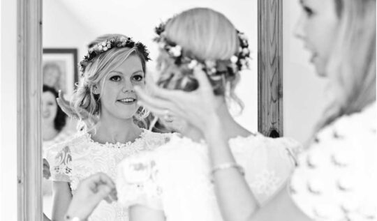 A bride with a soft, romantic updo adorned with a floral crown, getting ready for her wedding day in front of a mirror, showcasing the personalised bridal hair and makeup services by Jyn San Hair & Makeup in Somerset.