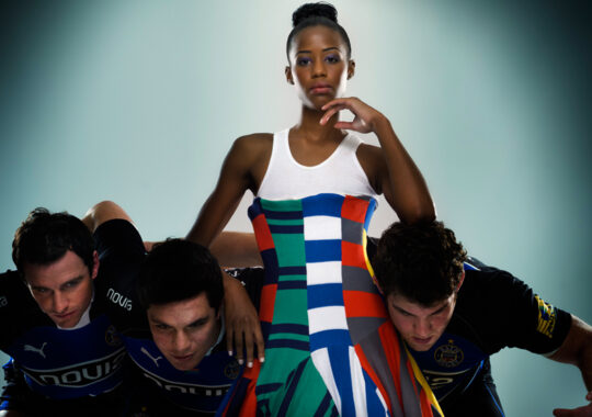 A bold fashion concept featuring a confident model in a colourful geometric dress standing poised above three rugby players in a scrum formation. The dramatic contrast between athletic determination and avant-garde styling highlights versatility in commercial styling and creative direction, blending sport and high fashion seamlessly