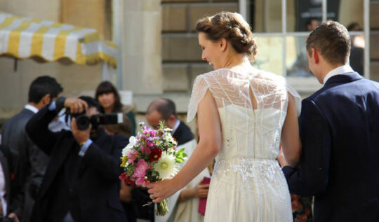 bride with a vintage-inspired updo and intricate beaded gown holding a vibrant bouquet, walking with her groom while being photographed, highlighting Jyn San Hair & Makeup's expert bridal styling services in Somerset