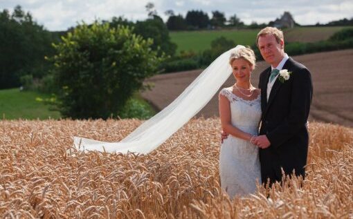 A bride with an elegant updo and natural makeup, wearing a lace wedding gown and flowing veil, standing with her groom in a picturesque wheat field, showcasing Jyn San Hair & Makeup's bridal styling services in Somerset.
