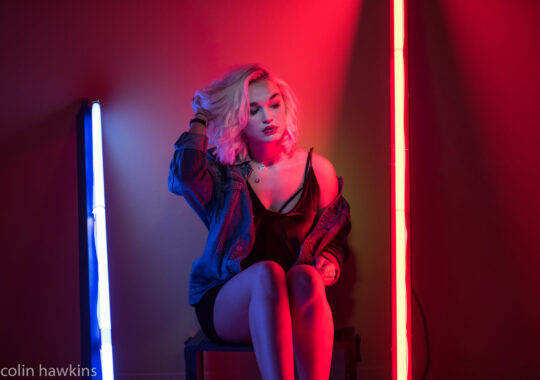 Edgy model illuminated by vibrant neon lighting, with a mix of red and blue tones creating a bold, modern aesthetic. She sports tousled platinum blonde hair, a denim jacket, and a black velvet dress with strappy detailing, perfect for commercial styling campaigns focused on nightlife, urban fashion, or alternative trends