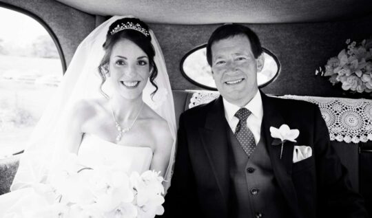 A radiant bride with an elegant curled updo and tiara, seated next to her father in a vintage car, showcasing Jyn San Hair & Makeup's professional bridal styling services in Somerset.