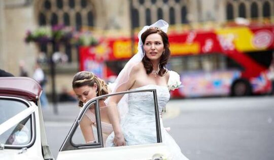 A bride with glamorous curls and natural makeup, stepping out of a classic wedding car with her bridesmaid assisting, showcasing the expert bridal hair and makeup services by Jyn San Hair & Makeup in Somerset.
