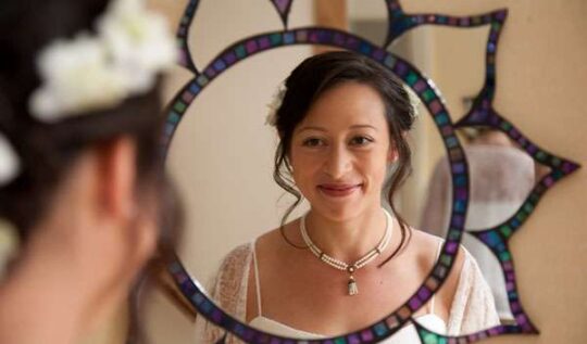 A bride with a soft, elegant updo and natural makeup, smiling into a decorative mirror while preparing for her wedding day, showcasing the bespoke bridal hair and makeup services by Jyn San Hair & Makeup in Somerset