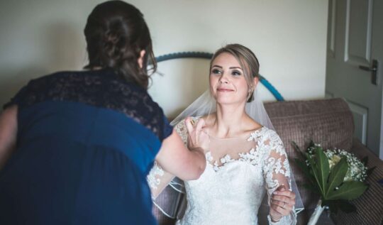 A bride with a sleek, elegant updo and natural makeup, receiving finishing touches as she prepares for her wedding day, showcasing the expert bridal hair and makeup services by Jyn San Hair & Makeup in Somerset.