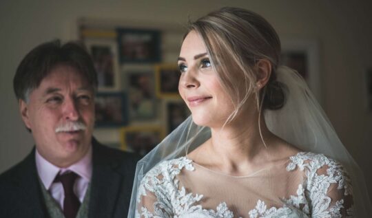A bride with a soft, elegant updo and radiant makeup, wearing a lace wedding gown and veil, smiling with her father by her side, showcasing Jyn San Hair & Makeup's bridal styling services in Somerset.