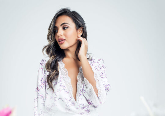 Elegant model with loose waves styled in a natural balayage, complemented by flawless makeup with soft pink lips and defined eyes. She wears a delicate floral robe with lace trim, posed against a minimal white background, highlighting luxury and sophistication for a commercial styling campaign