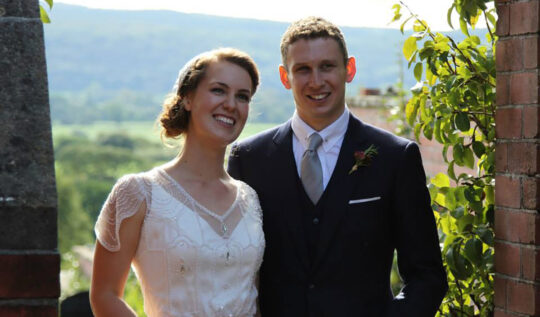 A bride with a vintage-inspired updo and soft makeup, standing with her groom in a scenic outdoor setting, showcasing Jyn San Hair & Makeup's expert bridal styling services in Somerset.