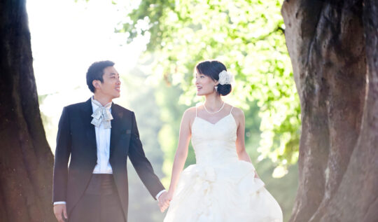 A bride with an elegant low bun adorned with a floral hairpiece and natural, radiant makeup, walking hand-in-hand with her groom in a sunlit garden, showcasing Jyn San Hair & Makeup's professional bridal styling services in Somerset.