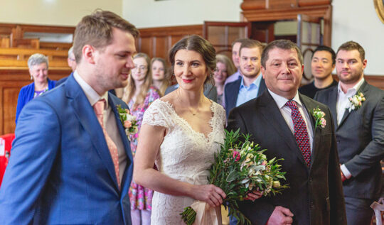 A bride with a classic low bun and natural makeup, walking down the aisle with her father in a beautifully lit ceremony, showcasing the expert bridal hair and makeup services by Jyn San Hair & Makeup in Somerset.