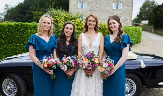 A bride with soft, natural makeup and flowing curls, standing with her bridesmaids in teal dresses holding vibrant bouquets, showcasing Jyn San Hair & Makeup's expert bridal and bridesmaid styling services in Somerset.