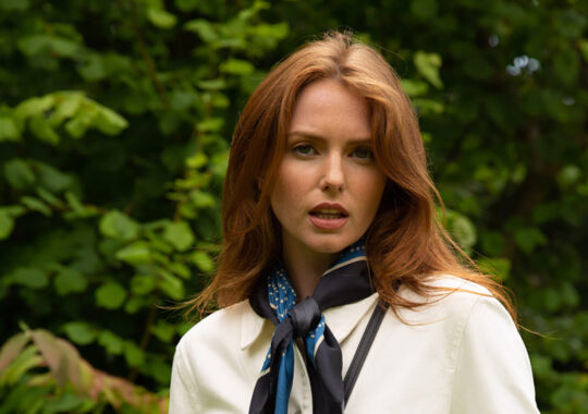Red-haired woman styled with natural makeup and soft waves, wearing a crisp white blouse paired with a navy silk scarf featuring polka dots. Set against a lush green outdoor background, the look exudes effortless sophistication and timeless elegance.